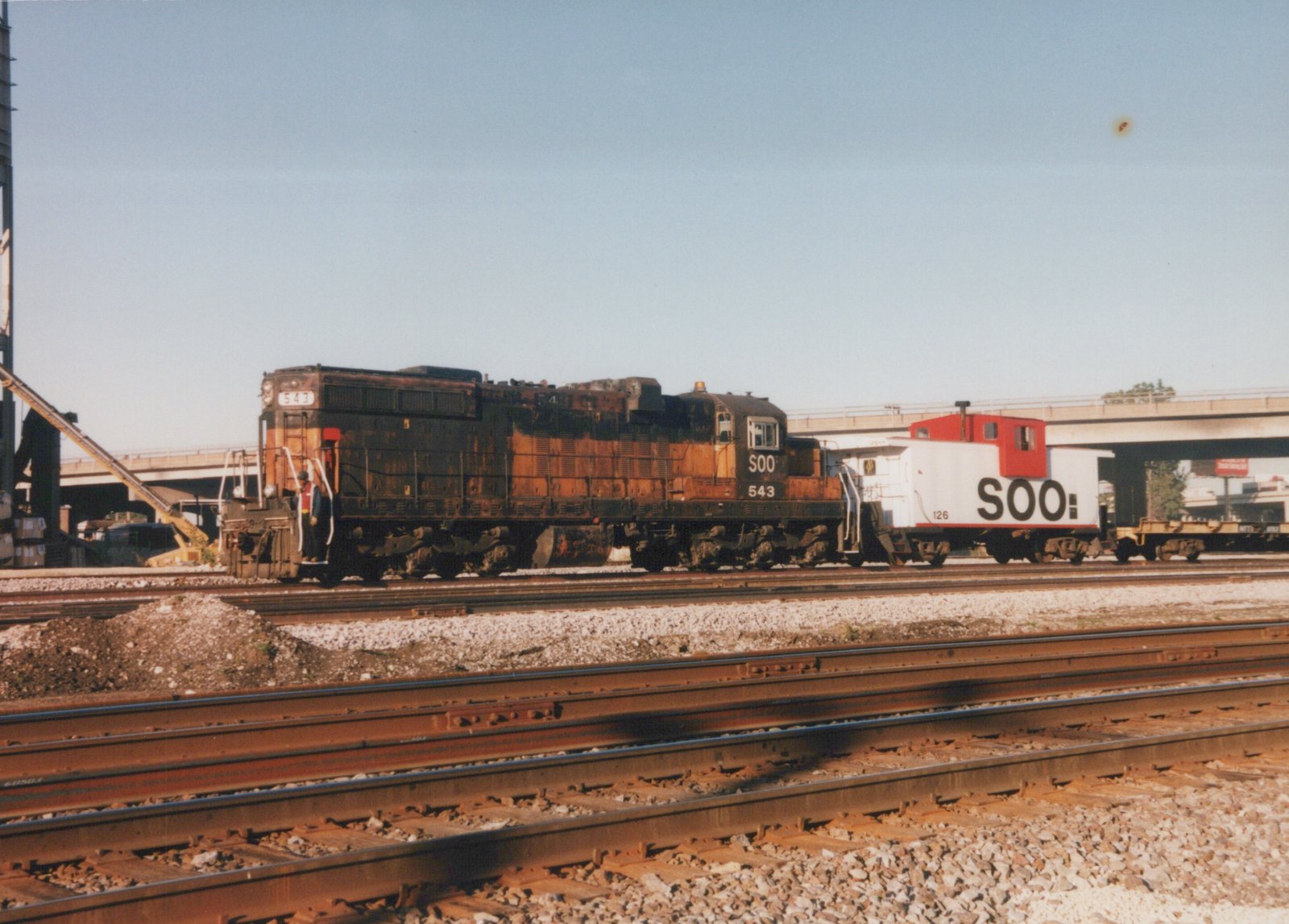 A rusty orange and black Soo Line diesel locomotive numbered 543 stands on a set of tracks next to a white and red caboose numbered 126, with an overpass in the background.