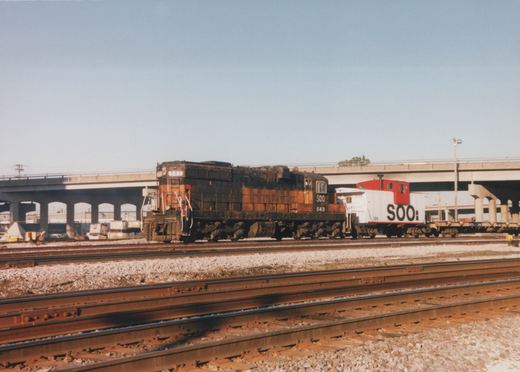 A rusty black and orange Soo Line diesel locomotive numbered 543 is coupled to a white and red Soo Line caboose numbered 126, stopped on multiple tracks in a rail yard with a large concrete bridge in the background under a clear blue sky.