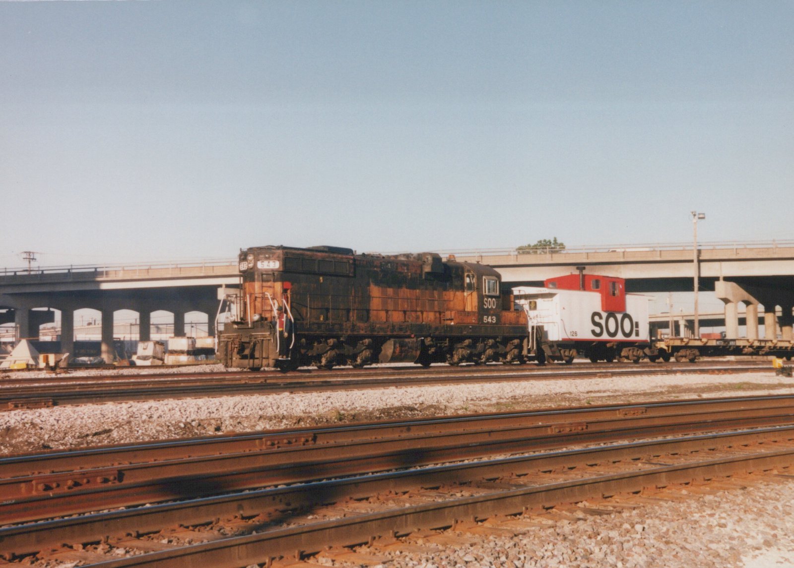 A rusty black and orange Soo Line diesel locomotive numbered 543 is coupled to a white and red Soo Line caboose numbered 126, stopped on multiple tracks in a rail yard with a large concrete bridge in the background under a clear blue sky.