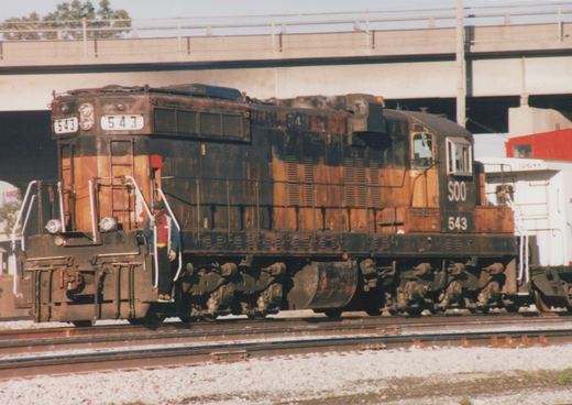 A rusty black and orange Soo Line diesel locomotive, numbered 543, is shown parked on tracks under a concrete bridge; a person in a cap and vest stands on its front platform.