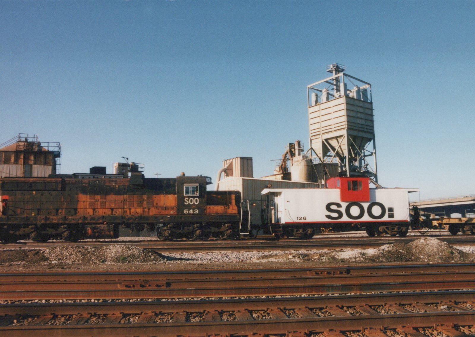 A Soo Line diesel locomotive and caboose parked on parallel tracks in a yard with industrial buildings in the background.