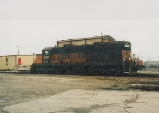 A side view of a black and orange Soo Line diesel locomotive numbered 543, parked on tracks in a gravel and concrete yard with overcast skies.