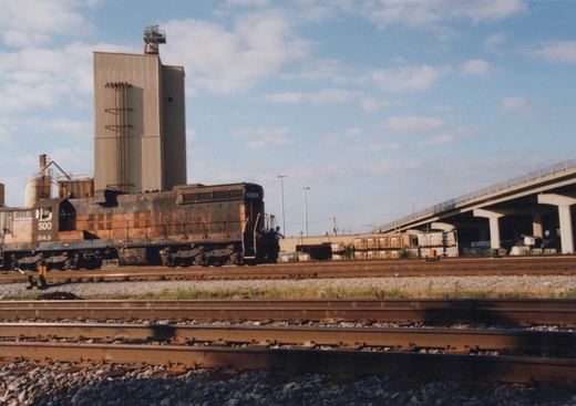 Side view of a rusty brown diesel locomotive with the number 500 and C&H lettering, parked on multiple tracks near an overpass and industrial buildings under a partly cloudy sky.