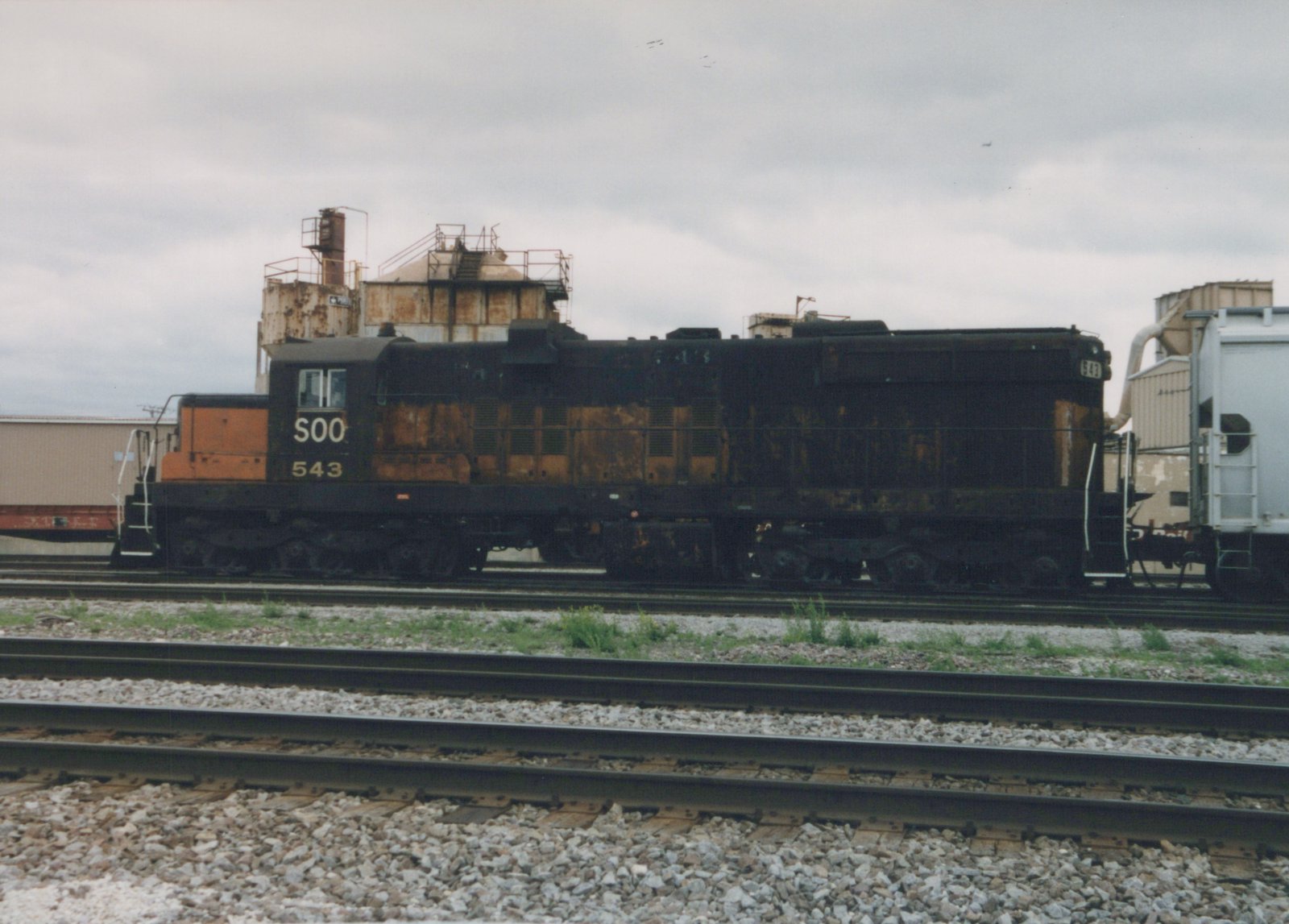 A black and orange Soo Line diesel locomotive numbered 543 is parked on tracks in a rail yard, with industrial buildings in the background under a cloudy sky.