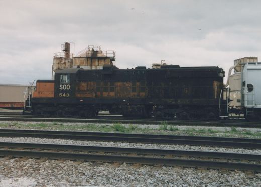 A black and orange Soo Line diesel locomotive numbered 543 is parked on tracks in a rail yard, with industrial buildings in the background under a cloudy sky.