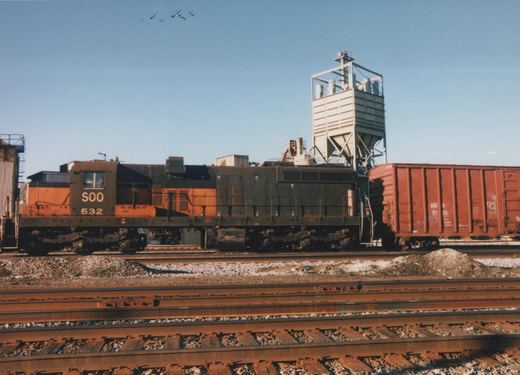 A side view of a dark gray and orange Soo Line diesel locomotive, number 532, standing on tracks in a yard with a red freight car behind it.