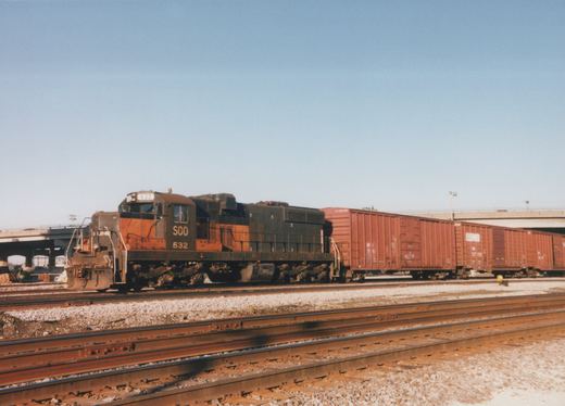 Side view of a black and orange Soo Line diesel locomotive numbered 532, pulling red boxcars, on a sunny day with multiple tracks and an overpass in the background.