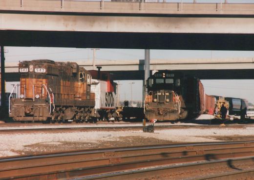 Two SOO Line diesel locomotives, number 543 in orange and black and number 517 in black with orange trim, are positioned on separate tracks under a concrete overpass, with a line of freight cars and a yellow excavator visible in the background.