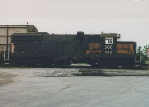 A side view of a black SOO Line diesel locomotive, number 532, sitting on tracks in a gravel yard with a building in the background under a gray sky.