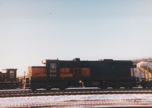 Side view of two black and orange Soo Line diesel locomotives on snow-covered tracks in a rail yard, clear blue sky.
