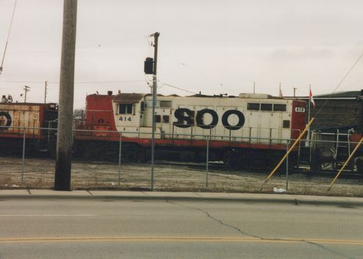A white and red SOO Line locomotive numbered 414 is visible behind a chain-link fence, with a freight car and industrial building in the background under an overcast sky.