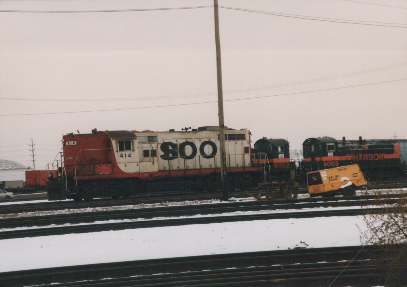 A snowy railroad yard featuring three diesel locomotives: SOO Line #414 and #9002, and Harbor Belt Railroad #9003, with a yellow maintenance railcar parked on an adjacent track.