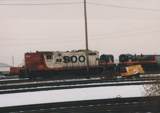 A snowy railroad yard featuring three diesel locomotives: SOO Line #414 and #9002, and Harbor Belt Railroad #9003, with a yellow maintenance railcar parked on an adjacent track.