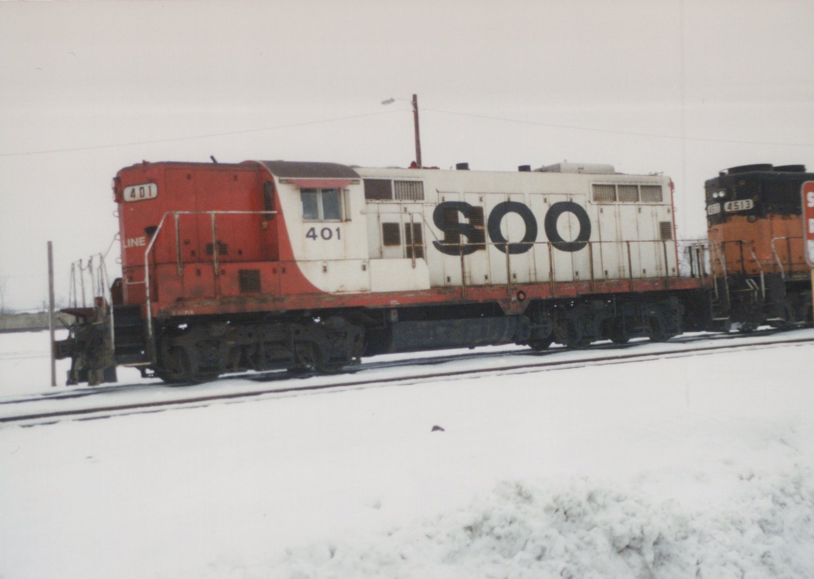 Two diesel locomotives, one red and white with the number 401 and 'SOO' lettering, and another orange and black with the number 4513, are parked side by side on snow-covered tracks.