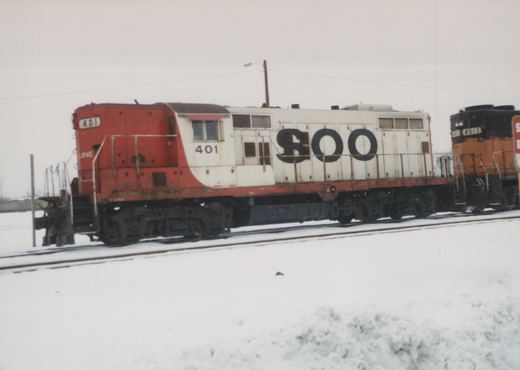 Two diesel locomotives, one red and white with the number 401 and 'SOO' lettering, and another orange and black with the number 4513, are parked side by side on snow-covered tracks.