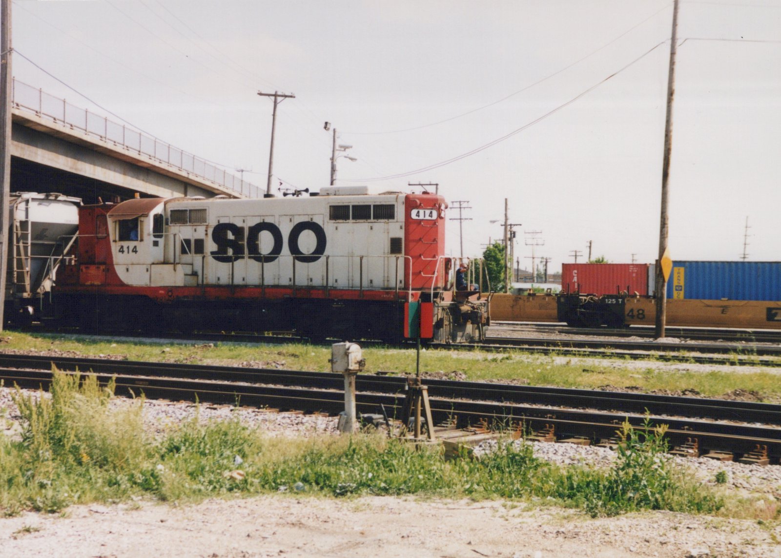 A white and red SOO Line diesel locomotive, numbered 414, stands on the tracks under a concrete overpass, with a person visible on the rear platform and intermodal containers on flatcars nearby.