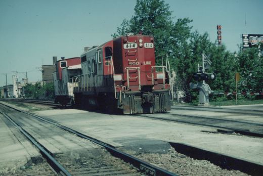 A red and grey SOO Line diesel locomotive numbered 414 is shown at a grade crossing in a suburban area with trees and a "MONT" sign visible.