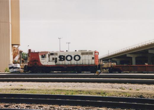 Side view of a white and red Soo Line diesel locomotive numbered 401, coupled to a red freight car, situated in a multi-track rail yard with a concrete overpass in the background.