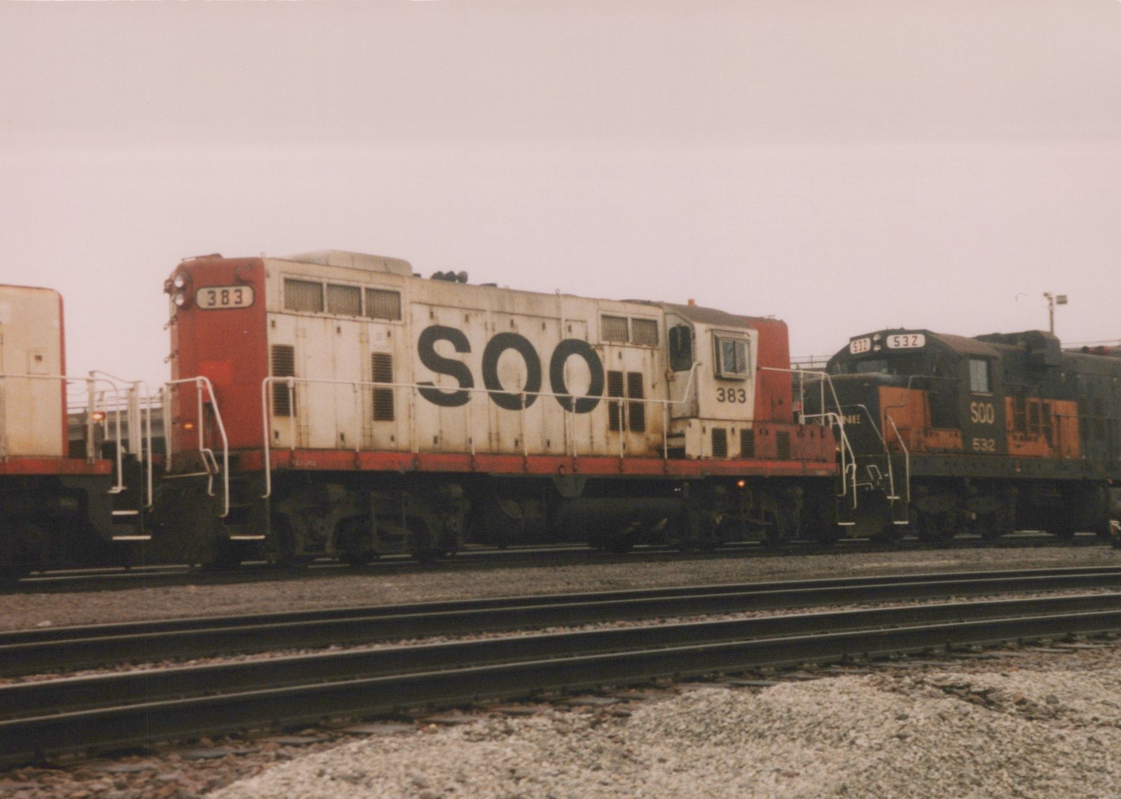Two diesel locomotives, one white and red, the other black and orange, both with 'SOO' lettering and distinct numbers, are parked on parallel railroad tracks under an overcast sky.