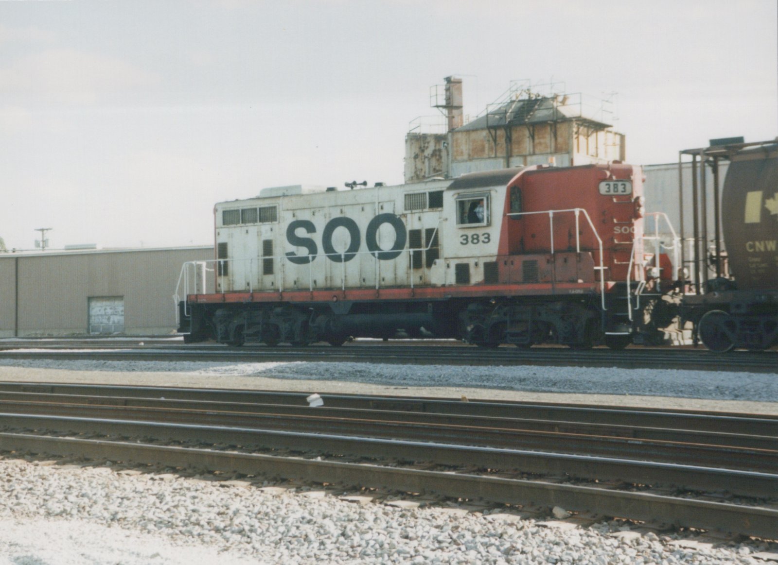 A white and red Soo Line diesel locomotive numbered 383 sits on a set of tracks in a yard, with a freight car labeled CNW visible to the right.