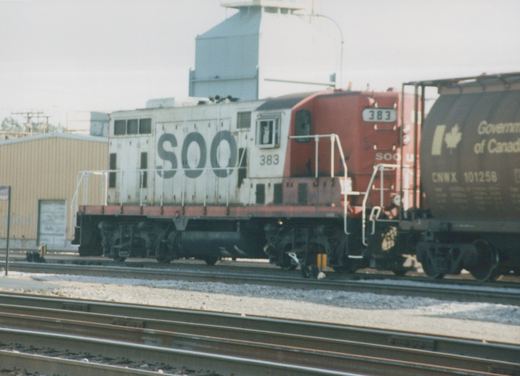 A side view of a white and orange SOO Line diesel locomotive, number 383, coupled to a dark brown Canadian National freight car in a rail yard with multiple tracks and a warehouse in the background.
