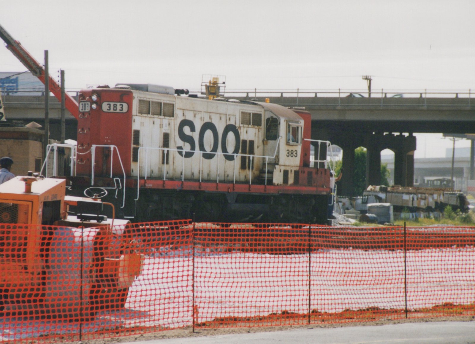 A white and red SOO Line diesel locomotive, number 383, is parked next to orange safety fencing with a bridge and construction equipment in the background.