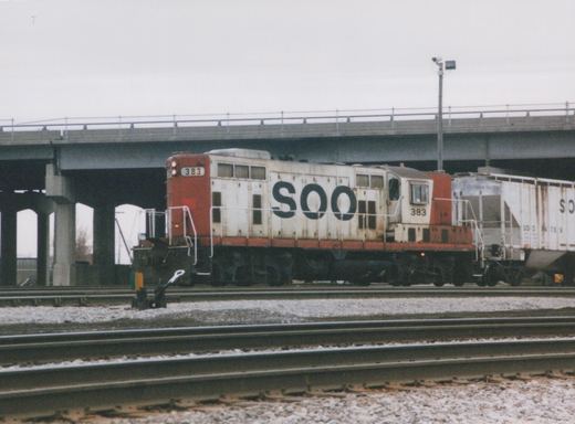 A side view of a red and white SOO Line diesel locomotive numbered 383 on railroad tracks, with a concrete bridge overhead and a freight car behind it.