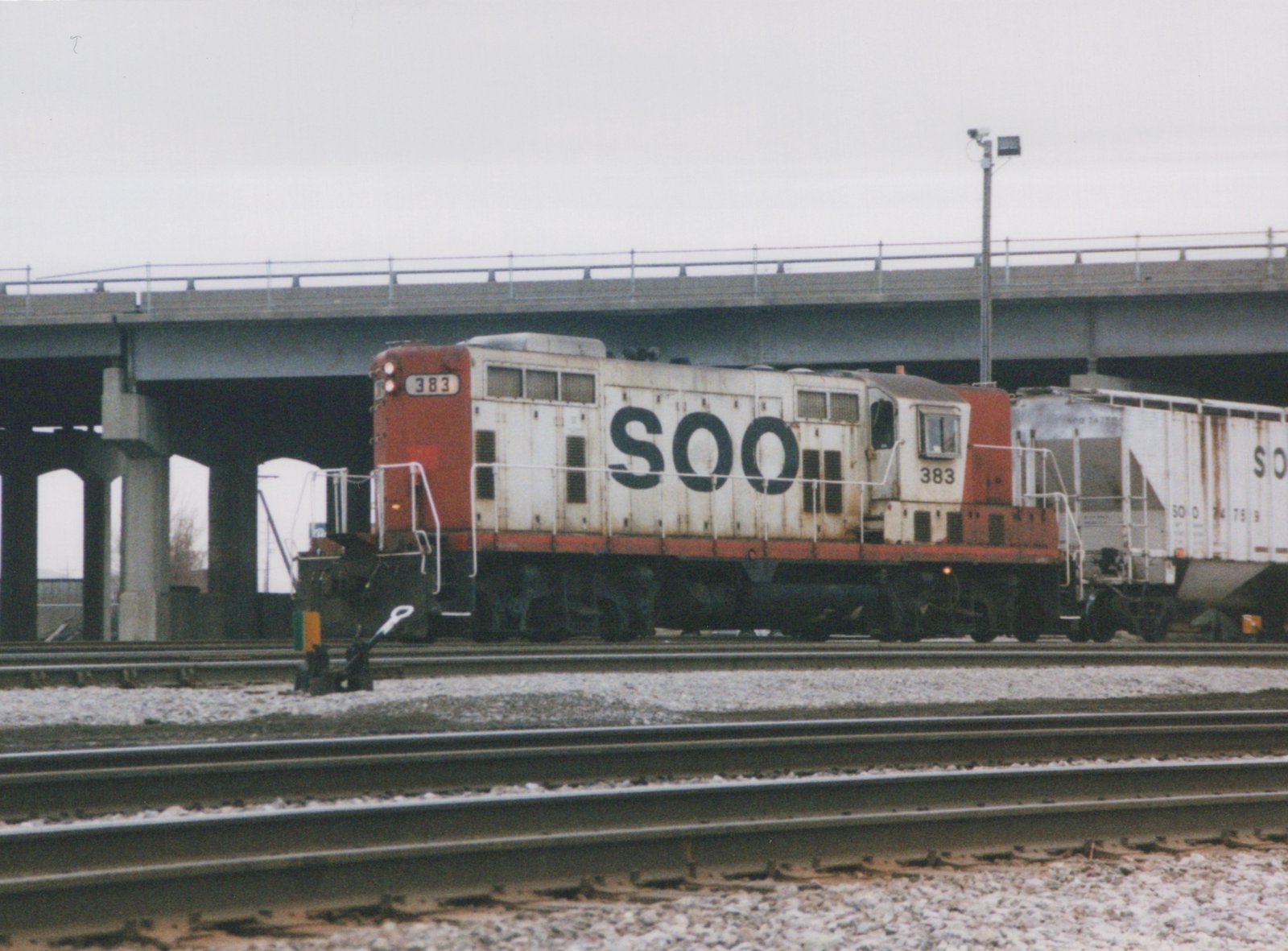 A side view of a red and white SOO Line diesel locomotive numbered 383 on railroad tracks, with a concrete bridge overhead and a freight car behind it.