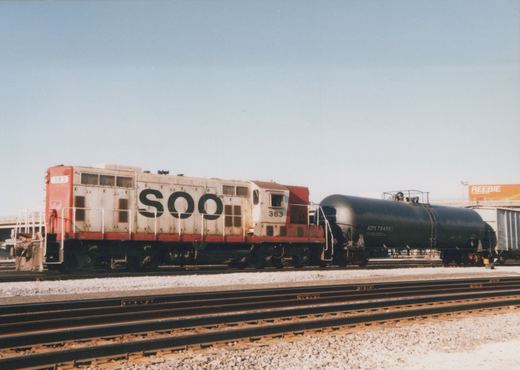 Side view of a white and red Soo Line diesel locomotive, number 383, coupled to a black tank car in a rail yard with multiple tracks.