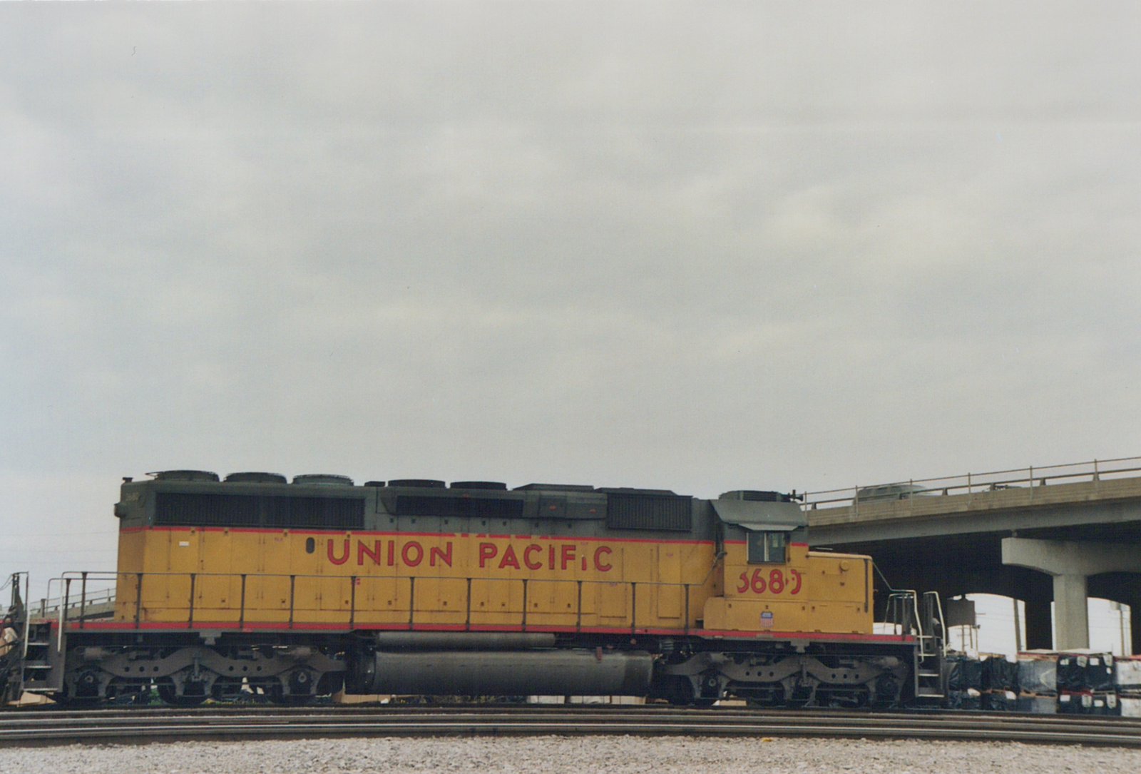 A yellow and gray Union Pacific diesel locomotive, number 3683, is parked on tracks under a concrete highway overpass on a cloudy day.