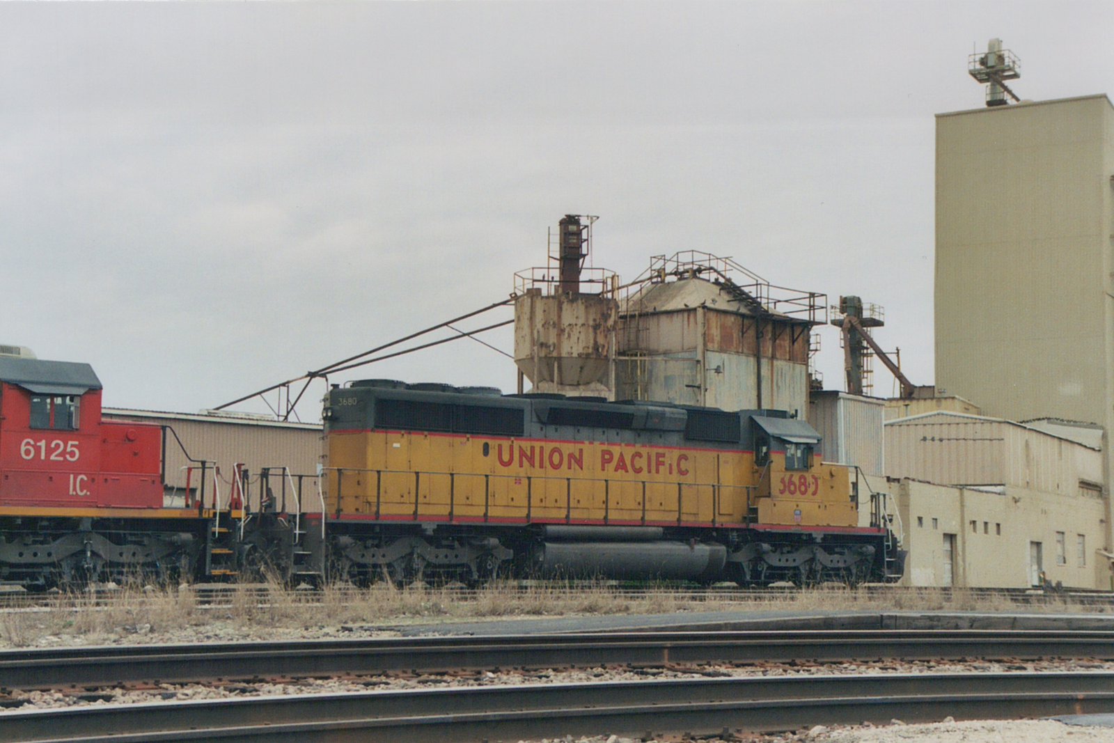 A yellow Union Pacific locomotive numbered 3680 and a red Illinois Central locomotive numbered 6125 sit on parallel tracks in an industrial yard under an overcast sky.