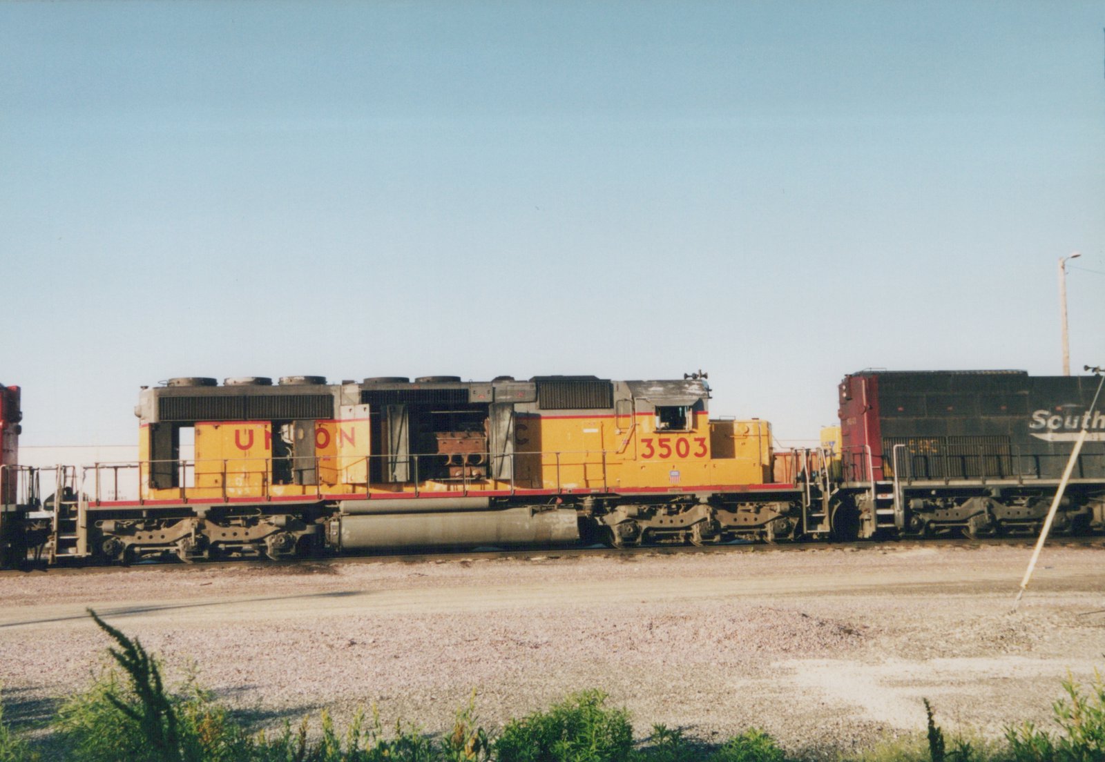 Side view of a yellow Union Pacific diesel locomotive numbered 3503, with a black Southern Railway locomotive partially visible beside it, parked on gravel in a sunny yard.