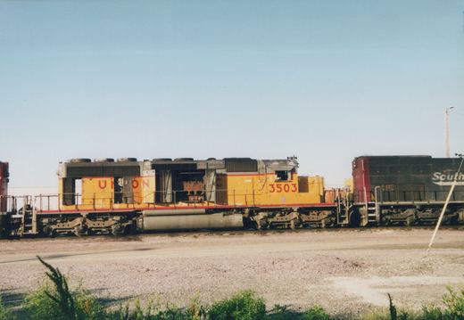 Side view of a yellow Union Pacific diesel locomotive numbered 3503, with a black Southern Railway locomotive partially visible beside it, parked on gravel in a sunny yard.