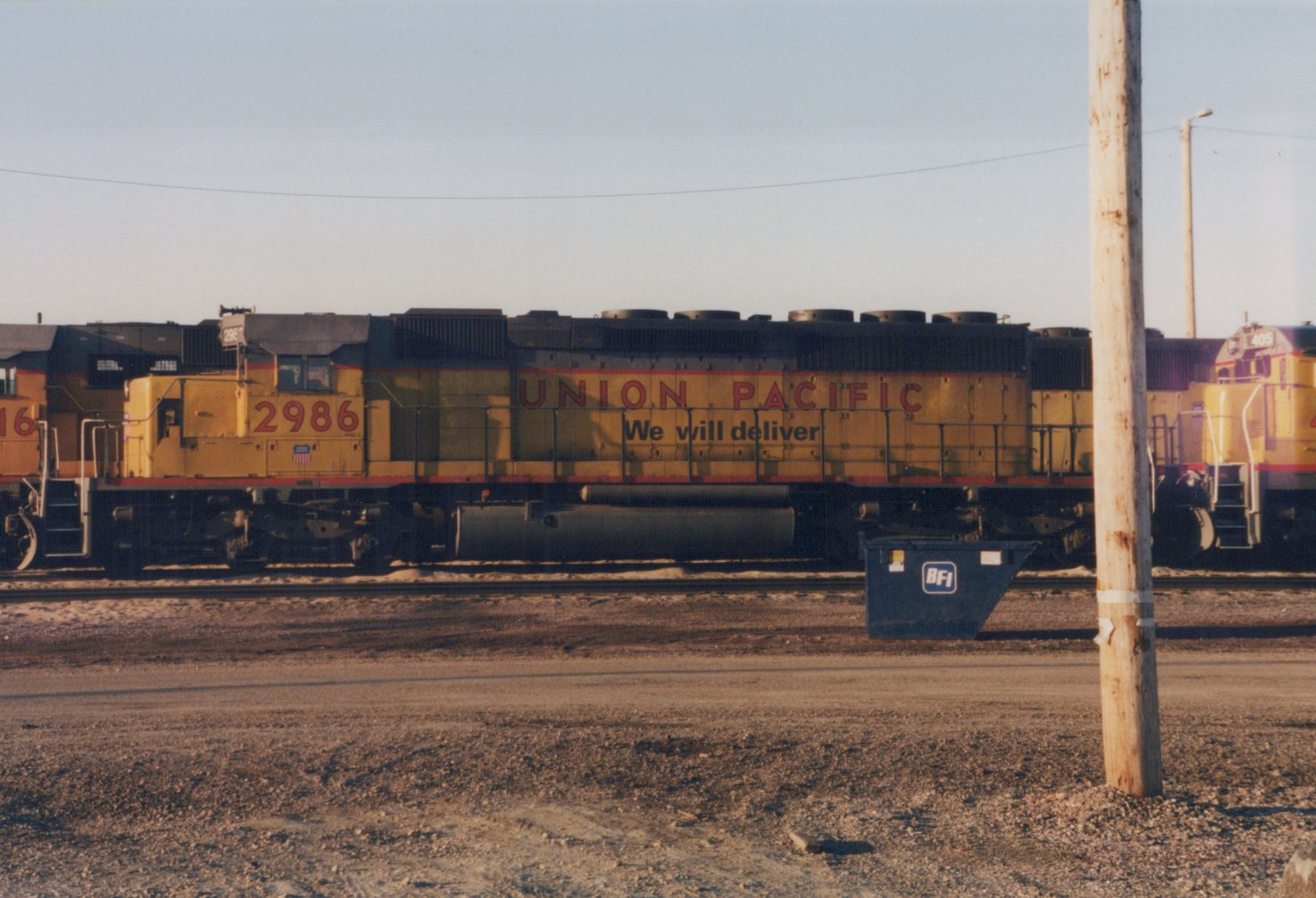 A side view of a yellow Union Pacific diesel locomotive numbered 2986, with another locomotive numbered 405 in the background, in a gravel yard with a blue dumpster and a utility pole nearby.