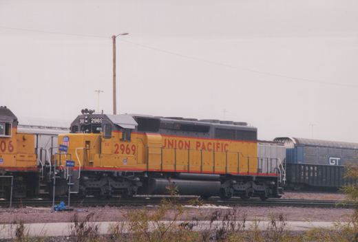 Side view of a yellow and gray Union Pacific locomotive number 2969 in a rail yard, with other trains and freight cars visible in the background under an overcast sky.