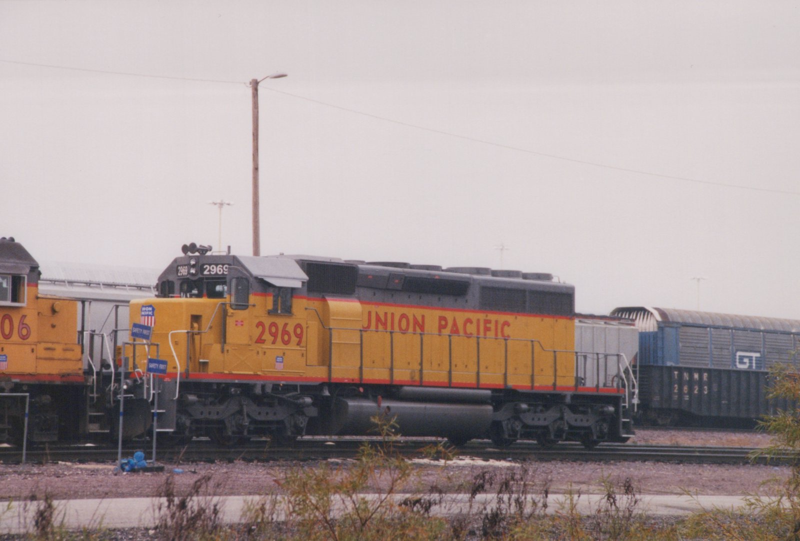 Side view of a yellow and gray Union Pacific locomotive number 2969 in a rail yard, with other trains and freight cars visible in the background under an overcast sky.