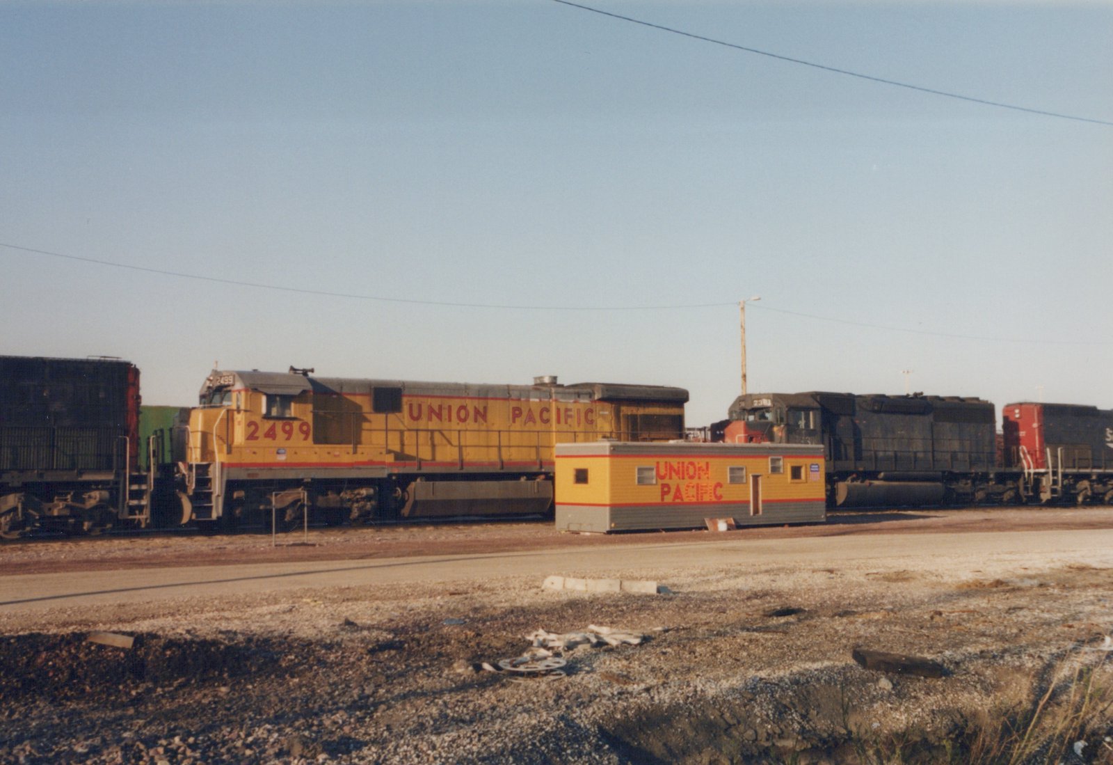 A yellow Union Pacific locomotive numbered 2499 sits in a gravel yard next to a dark-colored locomotive and a small yellow and gray office building, under a clear blue sky.