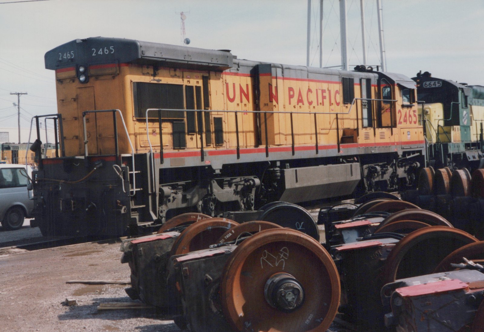 A yellow Union Pacific diesel locomotive numbered 2465 is parked in a yard, with a pile of old train wheels and trucks in the foreground and another locomotive visible behind it.