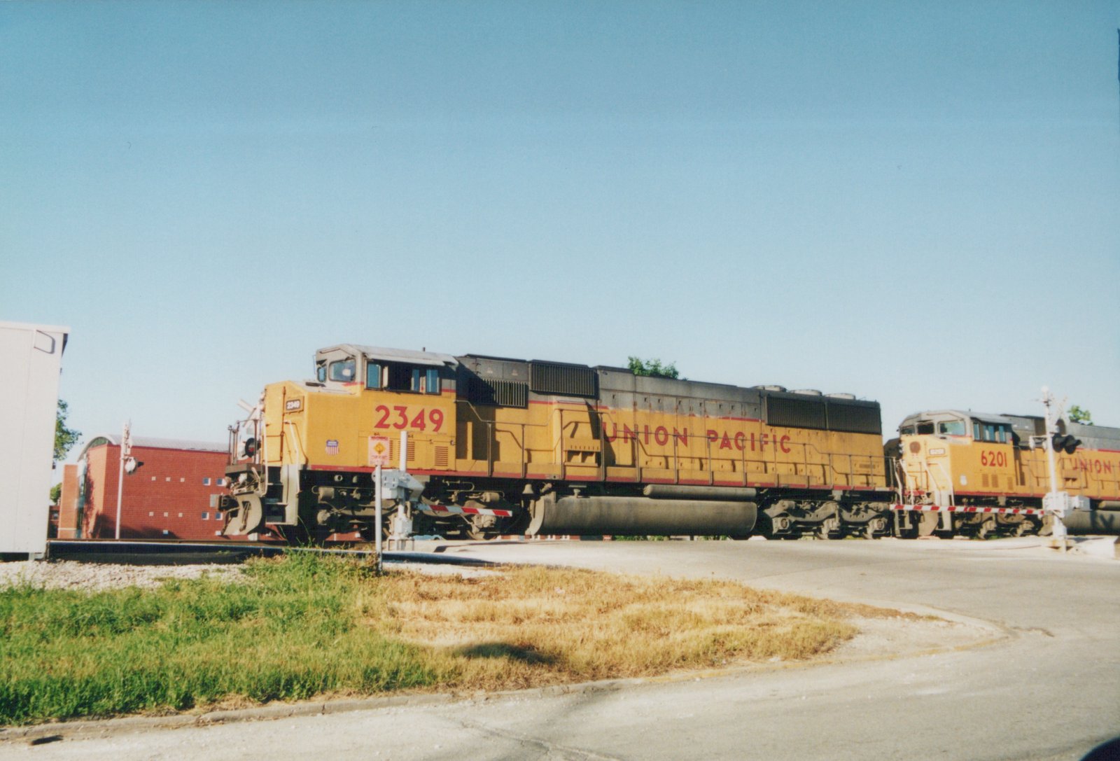 A side view of two yellow Union Pacific diesel locomotives at a railroad crossing under a clear blue sky with a small red brick building in the background.