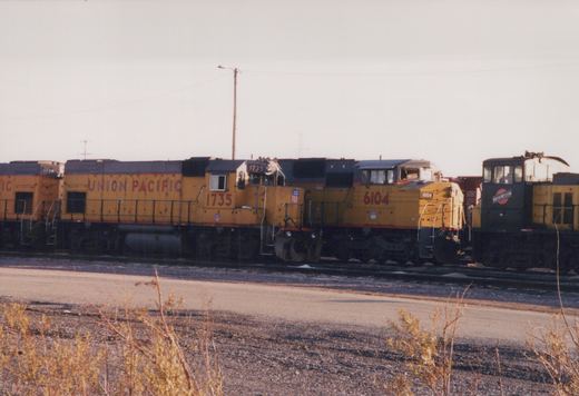 Three diesel locomotives are parked on parallel tracks in a gravel yard under a clear sky. The lead yellow locomotive is Union Pacific #1735, followed by Union Pacific #6104, and a dark blue Chicago and North Western locomotive is visible behind them.