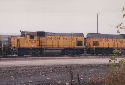 Two yellow and red Union Pacific diesel locomotives, numbered 1703 and 1714, sit side-by-side on a railroad track in a gravel yard with sparse vegetation and an overcast sky.