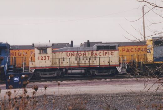 A side view of a Union Pacific diesel locomotive number 1237 in a rail yard on an overcast day with bare trees and other locomotives nearby.