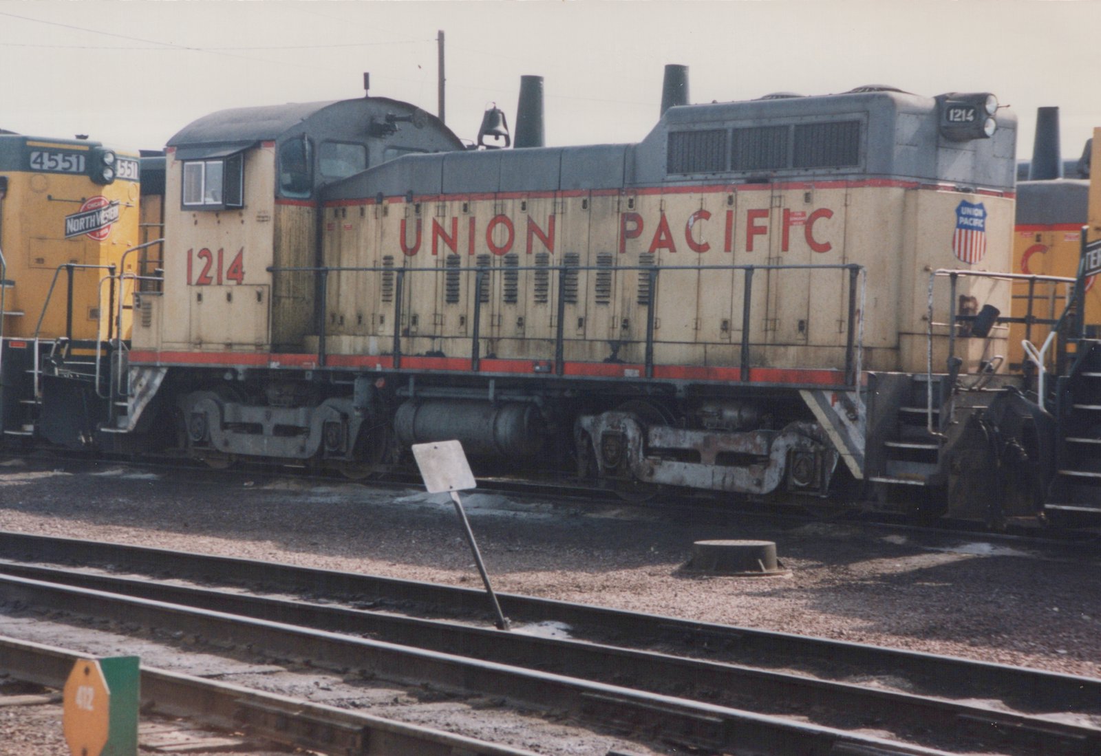 A side view of a Union Pacific SW1200 diesel locomotive, number 1214, parked in a rail yard with another locomotive visible behind it.