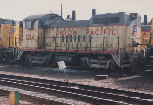 A side view of a Union Pacific SW1200 diesel locomotive, number 1214, parked in a rail yard with another locomotive visible behind it.