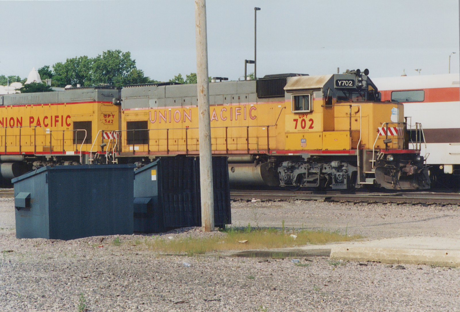 Side view of two yellow Union Pacific diesel locomotives numbered 702 and 547, with a white Amtrak passenger car in the background, parked on railroad tracks in a gravel yard.
