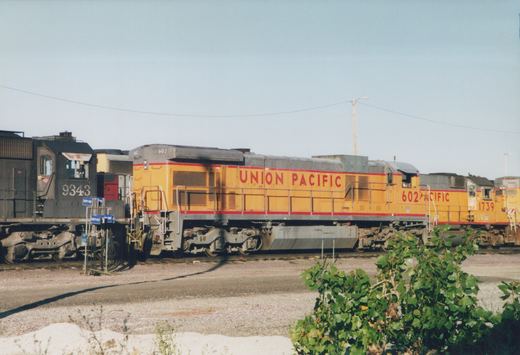 Three Union Pacific diesel locomotives in a gravel yard under a clear blue sky, with green bushes in the foreground.