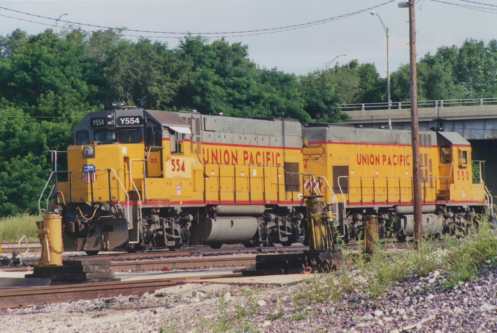 Two yellow Union Pacific locomotives, numbered Y554 and 550, are parked side-by-side on a railroad track in a yard, with trees and an overpass in the background.