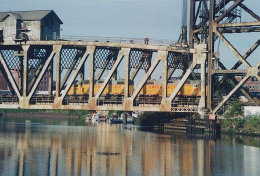 Two yellow and gray Union Pacific diesel locomotives numbered 418 and 419 are seen crossing a large metal truss bridge over a river with boats moored below.