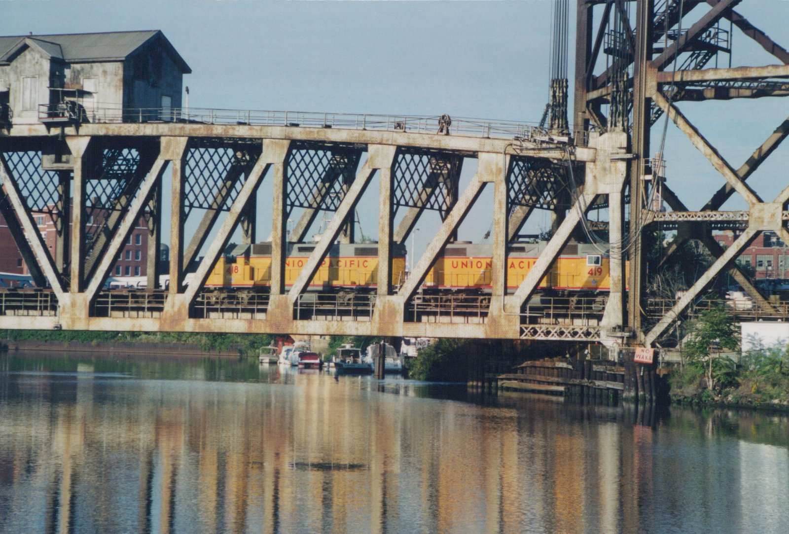Two yellow and gray Union Pacific diesel locomotives numbered 418 and 419 are seen crossing a large metal truss bridge over a river with boats moored below.