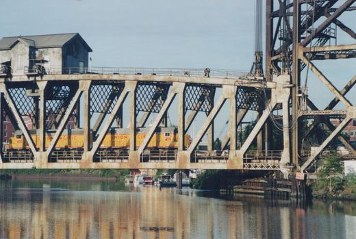 Two yellow and gray Union Pacific E9 locomotives numbered 418 and 419 travel across a steel truss bridge over a river, with buildings in the background and boats moored below.